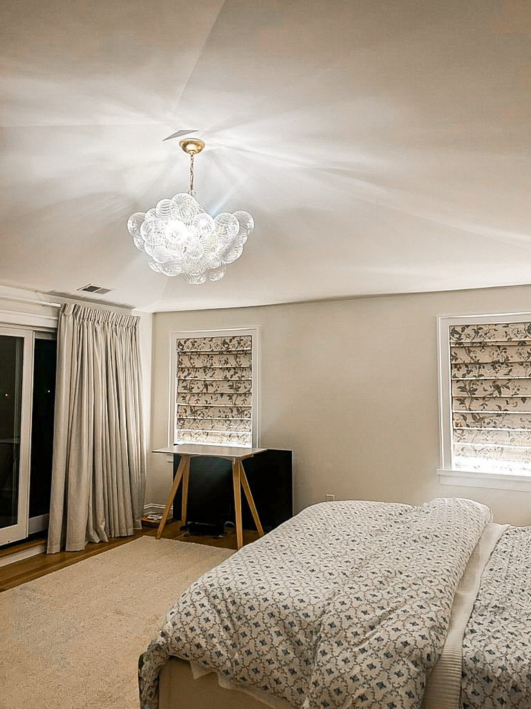 Primary bedroom in a Branford Connecticut new build with a ribbed glass bubble chandelier, botanical roman shades, and neutral linen curtains — interior design by Seawillow Studio