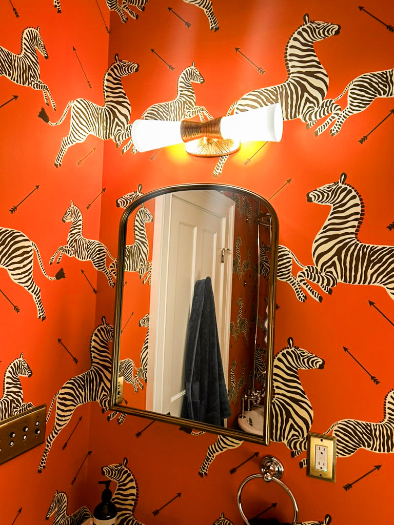 Guest bathroom featuring bold orange zebra wallpaper, an arched brass mirror, and a gold double sconce — coastal Connecticut interior design by Seawillow Studio Milford CT
